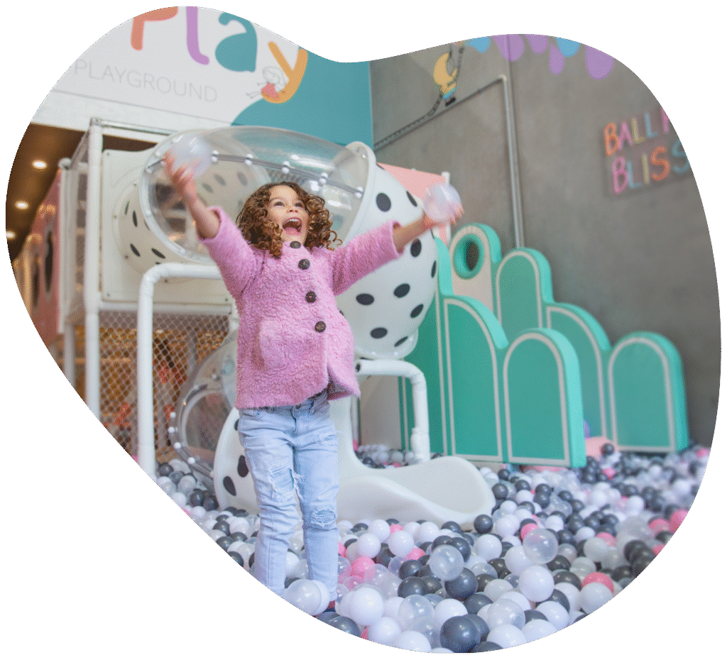 A young girl with curly hair joyfully playing in a ball pit, holding clear balls with her arms raised at Pixie Play Cafe
