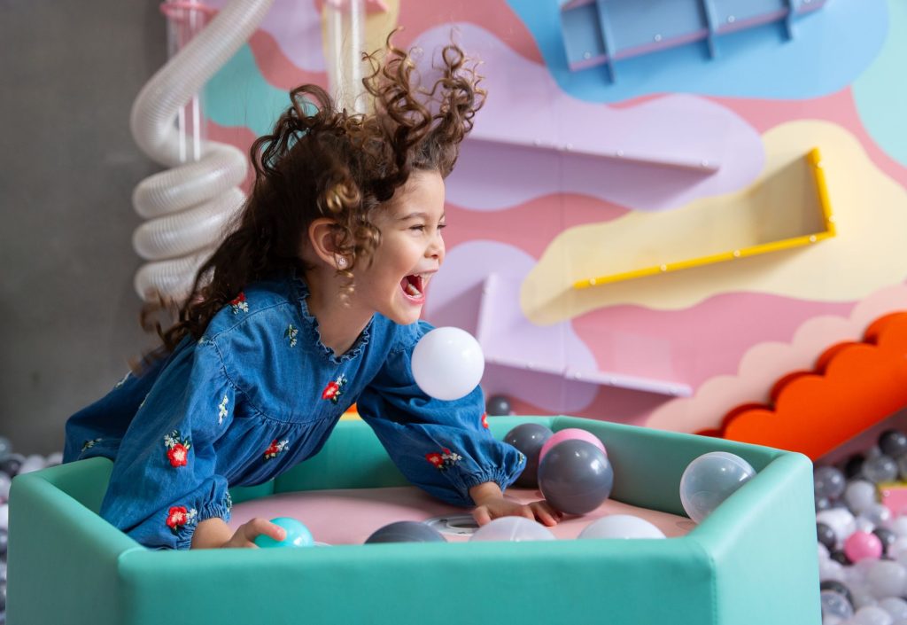 A joyful girl in a blue denim dress with red floral embroidery leans over a low, padded teal structure.
