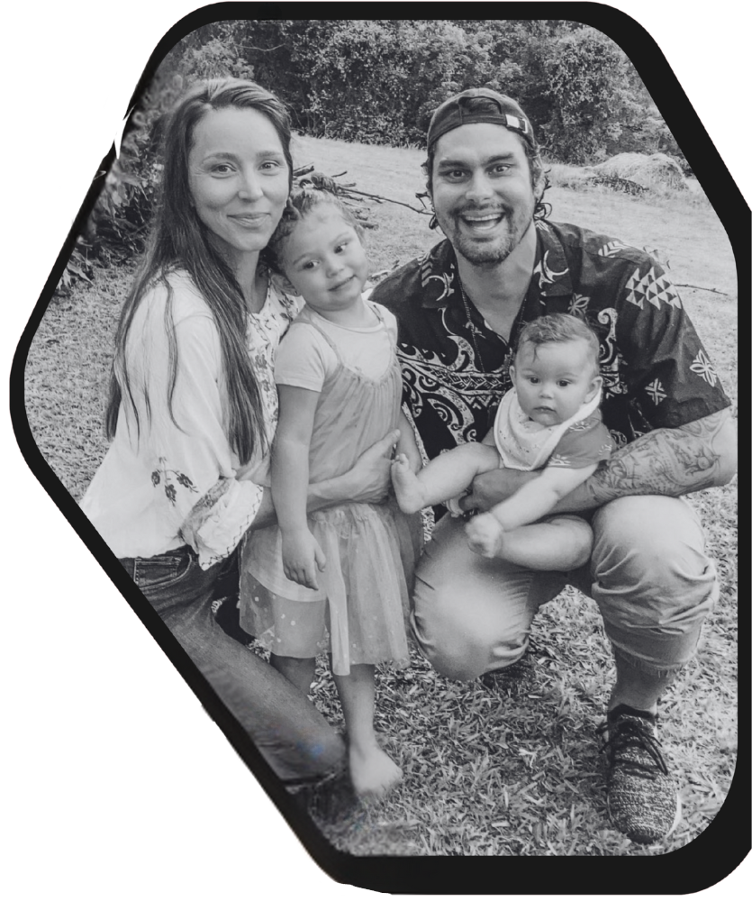 A family of four posed closely together in a casual, outdoor setting, framed within a decorative hexagonal border.