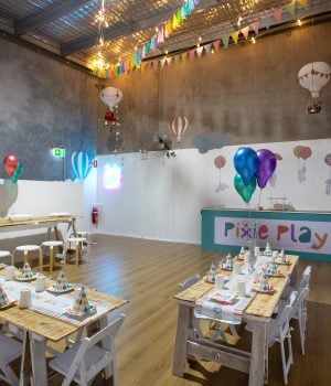 A wide shot of a large room set up for a child's birthday party, decorated with hot air balloons, string lights, and colorful bunting.