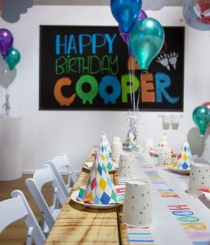 A close-up of a wooden party table set with colorful, diamond-patterned birthday hats and gold-dotted cups, in front of a chalkboard sign reading "HAPPY BIRTHDAY COOPER."
