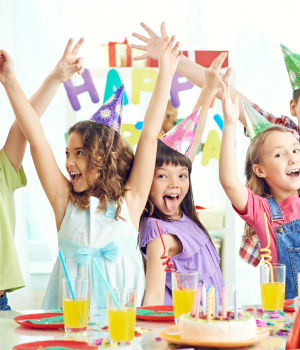 A group of five excited children wearing party hats, cheering with their hands in the air over a table set for a birthday party.